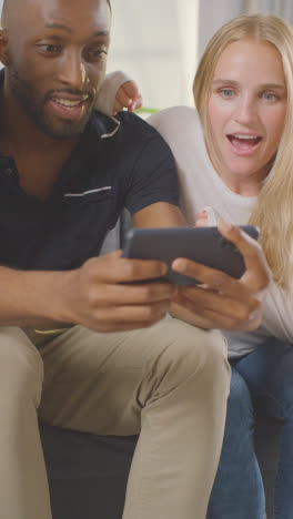 Vertical-Video-Of-Multi-Cultural-Friends-Celebrating-Watching-Sports-Game-On-Mobile-Phone-Sitting-On-Sofa-At-Home-